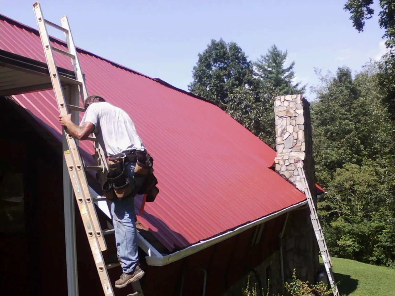 Roofer on a ladder beside a red metal roof and stone chimney — Soffit & Fascia Repair by Roofing Brothers in Litchville